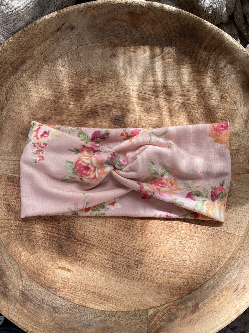 May include: A pink floral headband with a twisted knot design. The headband features a variety of pink, yellow, and green floral patterns.