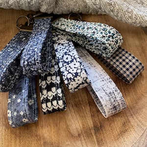 May include: A set of six keychains with different fabric patterns. The keychains are made of fabric with various floral and geometric designs in shades of blue, white, and black. The keychains are arranged on a wooden surface.