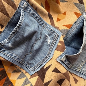 May include: Two blue denim jean pockets are placed on a patterned fabric. The pockets are a medium wash denim with visible stitching and distressed details. The fabric beneath has a geometric design in shades of brown, tan, and orange.