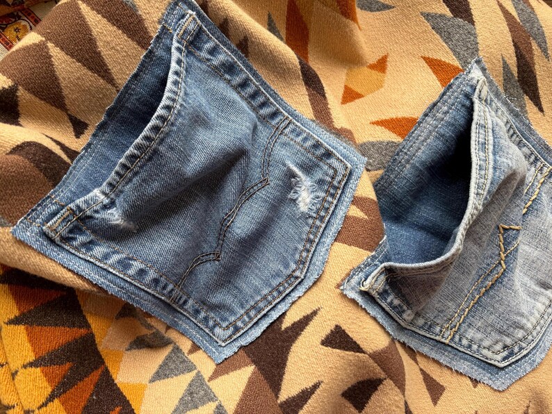 May include: Two blue denim jean pockets are placed on a patterned fabric. The pockets are a medium wash denim with visible stitching and distressed details. The fabric beneath has a geometric design in shades of brown, tan, and orange.