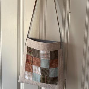 May include: A beige fabric crossbody bag featuring a central square panel made of a patchwork of plaid and checkered fabric squares in shades of brown, tan, white, and light blue. The bag has a long, thin, matching fabric shoulder strap.
