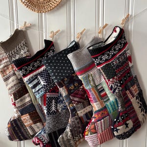 May include: A collection of six Christmas stockings, each with a unique patchwork design. The stockings are made of various fabrics and patterns, including snowflakes, stars, and geometric shapes. They are hung on a white wall with wooden clothespins.