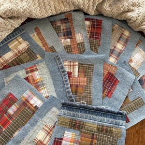 May include: A collection of square fabric patches featuring blue denim borders and center panels of assorted plaid and checkered cotton textiles in shades of red, brown, tan, and white. The patches have frayed edges and are layered together.