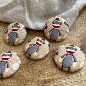 May include: Five round fabric buttons with a beige background and a pattern of gray monkeys wearing red hats. The monkeys are smiling and have red mouths.