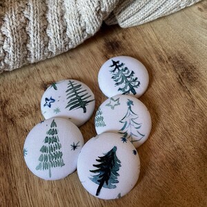 May include: Five white fabric-covered buttons with green and blue Christmas tree designs and star accents. The buttons are arranged on a wooden surface, with a portion of a beige knit sweater visible in the background.