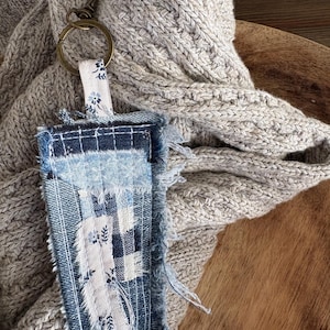 May include: A handmade denim keychain with a patchwork design. The keychain features a rectangular denim tag with a white floral pattern and a brass-colored ring. The background includes a gray knitted scarf and a wooden surface.