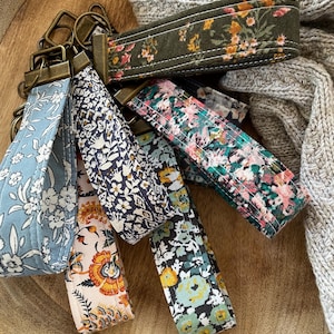 May include: A set of six colorful keychains with floral patterns. Each keychain is made of fabric and has a metal ring for attaching keys. The keychains are arranged in a wooden bowl.