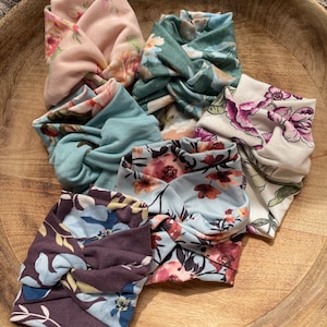 May include: Six floral print fabric headbands in a variety of colors and patterns, including pink, green, blue, and brown. The headbands are arranged in a circle on a wooden surface.