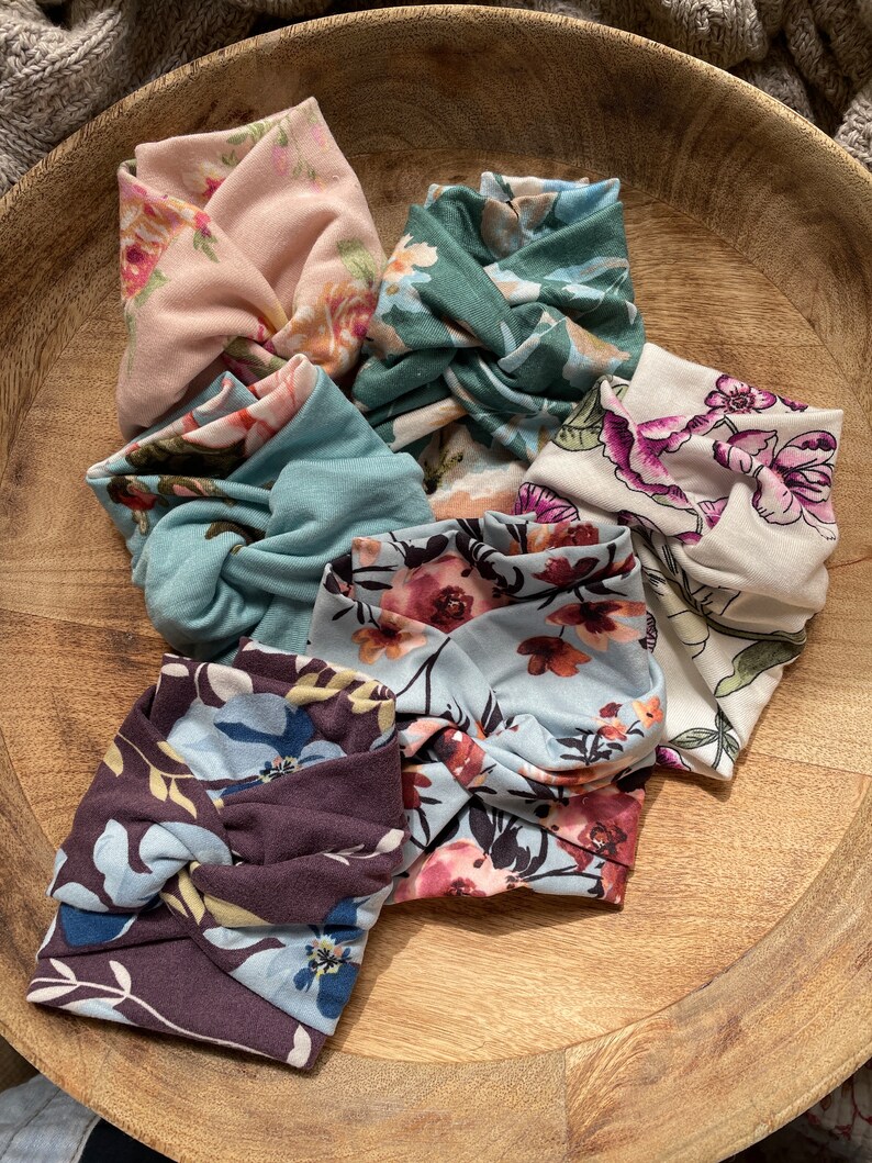 May include: Six floral print fabric headbands in a variety of colors and patterns, including pink, green, blue, and brown. The headbands are arranged in a circle on a wooden surface.