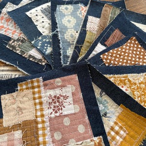 May include: A collection of square fabric patches featuring dark blue denim bases layered with various textile scraps. The pieces display diverse patterns including floral, polka dot, gingham, and geometric prints in shades of mustard, pink, and white, all secured with visible white running stitch quilting.