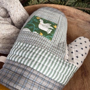 May include: A quilted oven mitt with a patchwork design. The mitt features a square patch with a white bird on a green background. The main body has a plaid pattern with green and white stripes. The cuff is yellow.