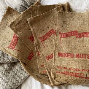 May include: Six small burlap bags with red printed flags and the words "Country Store" and "Mixed Nuts". The bags are stacked on top of each other and have a rustic, vintage look.