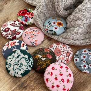 May include: Assortment of nine round fabric buttons with floral patterns. The buttons are in various colors, including pink, blue, green, and white. The buttons are arranged on a wooden surface.