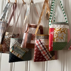 May include: A collection of five quilted crossbody bags hanging in a row. Each bag features a unique patchwork design made from various fabric scraps in shades of brown, green, red, and white, with long fabric straps and stitched patterns.
