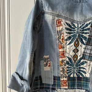 May include: Light blue denim jacket with patchwork details. The back features a large panel with a teal and white floral design, complemented by plaid and floral fabric patches. The jacket has rolled-up sleeves and a white lace trim along the bottom.