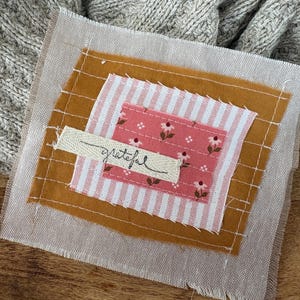 May include: A layered fabric patch featuring a light beige base with white grid stitching, a mustard yellow square, a pink and white striped rectangle, and a small pink floral square. A cream fabric strip with the word grateful is stitched on top.