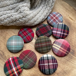 May include: Ten round fabric buttons with plaid patterns in red, green, blue, and white. The buttons are arranged on a wooden surface.