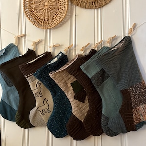 May include: A collection of eight Christmas stockings in various shades of brown, green, and blue, hanging from a string with wooden clothespins. Some stockings feature textured knit patterns and patches. Two woven wall decorations are above the stockings.