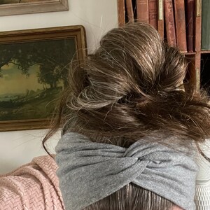 May include: A gray twisted headband made of soft fabric. The headband is worn by a person with long, brown hair pulled back in a bun.