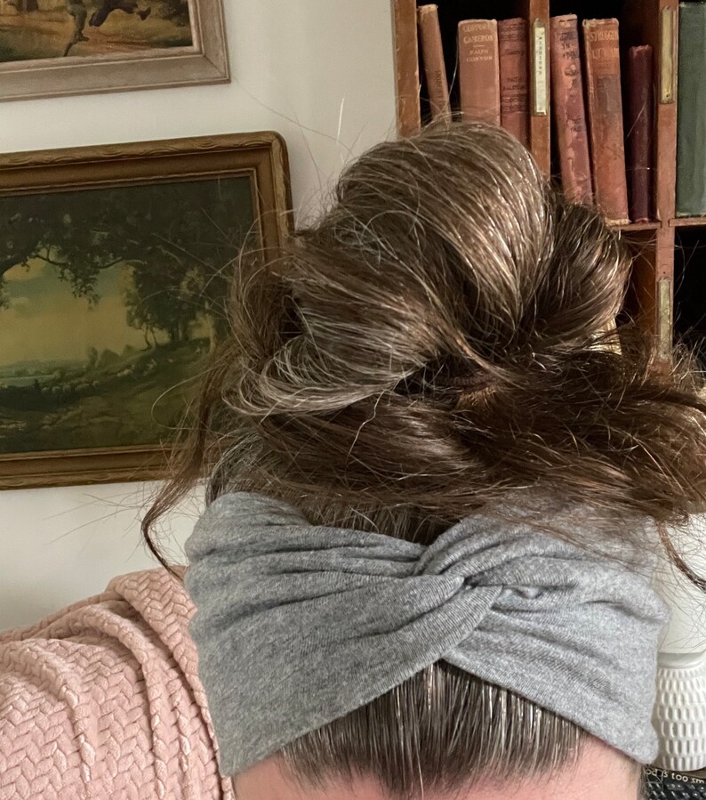 May include: A gray twisted headband made of soft fabric. The headband is worn by a person with long, brown hair pulled back in a bun.