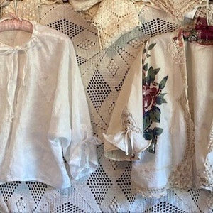 Puede incluir: Dos blusas blancas de lino con ribete de encaje. Una blusa tiene un escote fruncido y la otra tiene un estampado floral.