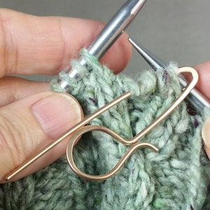 May include: Close-up of a green knitted fabric with two knitting needles and a gold-colored stitch holder. The needles are positioned to hold the stitches in place.