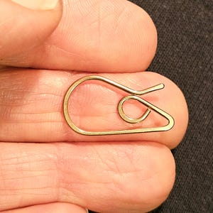 May include: A small, gold-colored paper clip with a unique, looped design. The clip is held between fingers, showcasing its intricate shape and metallic sheen. The design features a curved body and a spiral detail.