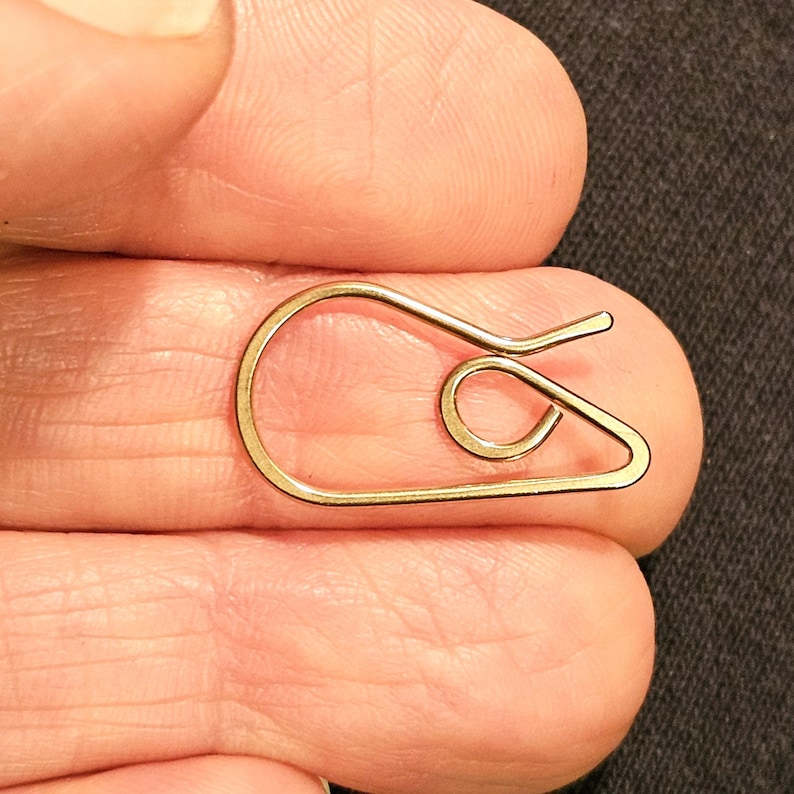 May include: A small, gold-colored paper clip with a unique, looped design. The clip is held between fingers, showcasing its intricate shape and metallic sheen. The design features a curved body and a spiral detail.