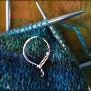 May include: Close-up of knitting needles with blue and green yarn in progress. A silver stitch marker is attached to the work. The yarn is a mix of blues and greens, creating a textured effect. The needles are silver and the background is a mix of colors.