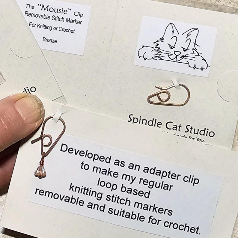 May include: Bronze "Mousie" clip removable stitch markers for knitting or crochet. The image shows the product on a card with the text "Spindle Cat Studio" and a cat illustration. The card also includes the text "Developed as an adapter clip to make my regular loop based knitting stitch markers removable and suitable for crochet."