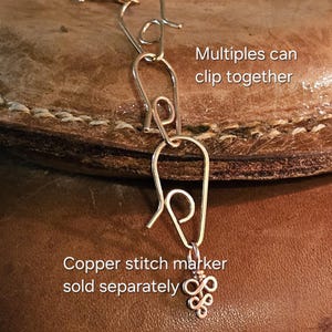 May include: Close-up of a silver-colored stitch marker with a decorative charm. The text "Multiples can clip together" and "Copper stitch marker sold separately" are visible. The marker is attached to a leather surface.