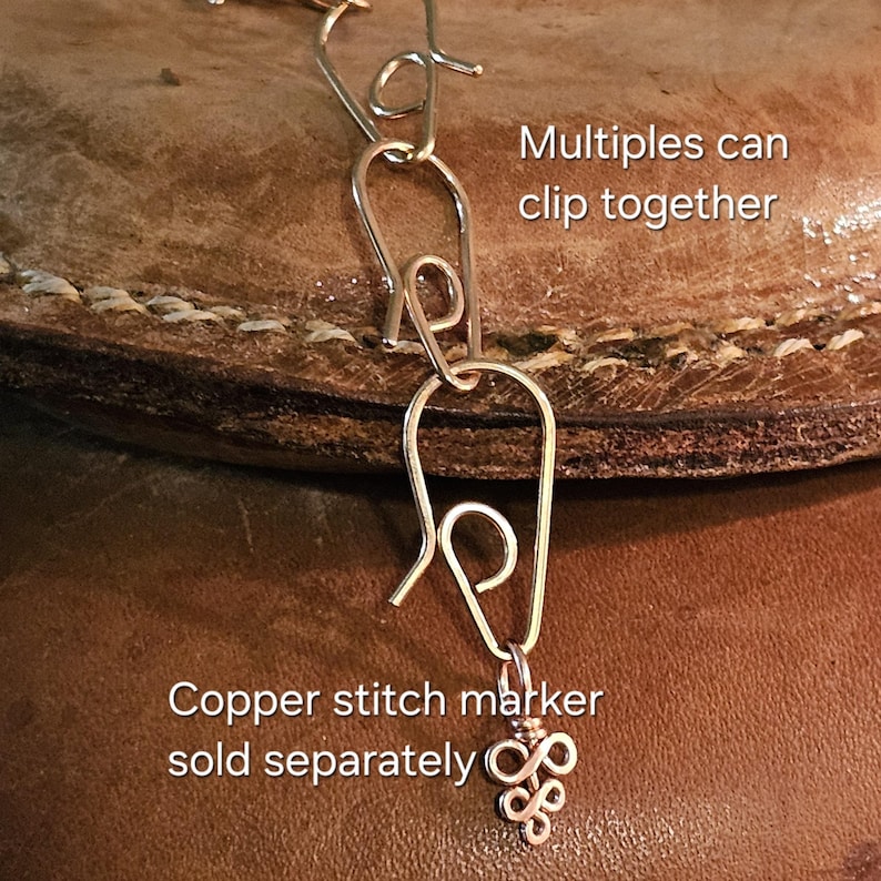 May include: Close-up of a silver-colored stitch marker with a decorative charm. The text "Multiples can clip together" and "Copper stitch marker sold separately" are visible. The marker is attached to a leather surface.