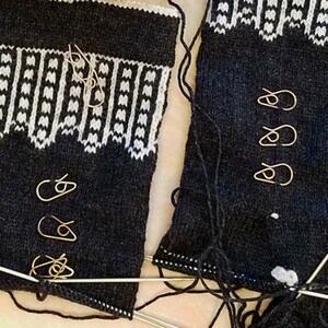 May include: Close-up of a knitting project in progress. Two sections of dark blue knitted fabric with a white geometric pattern at the top. Several gold-colored stitch markers are attached. Knitting needles and loose yarn are also visible.