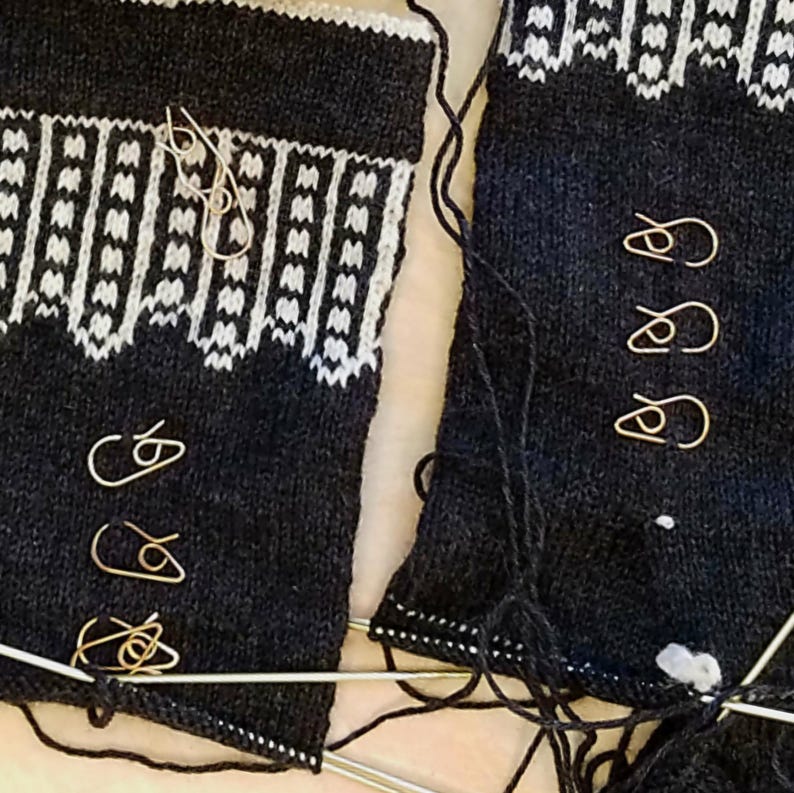 May include: Close-up of a knitting project in progress. Two sections of dark blue knitted fabric with a white geometric pattern at the top. Several gold-colored stitch markers are attached. Knitting needles and loose yarn are also visible.
