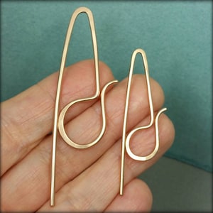 May include: Two gold-toned metal earrings with a minimalist wire design. Each earring features a long, straight vertical bar connected to a rounded, teardrop-shaped loop that curves outward at the bottom, creating a continuous, fluid silhouette.