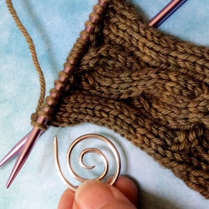 May include: A close-up of a green knitted fabric with a cable stitch pattern. A silver spiral stitch marker is in the foreground. Two purple knitting needles are visible in the background.