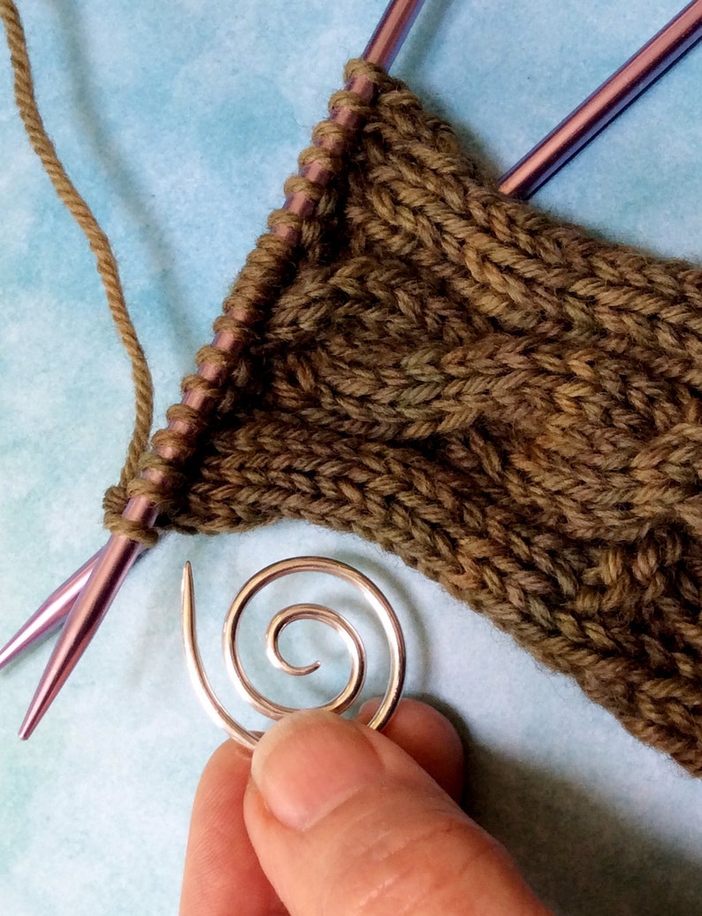 May include: A close-up of a green knitted fabric with a cable stitch pattern. A silver spiral stitch marker is in the foreground. Two purple knitting needles are visible in the background.