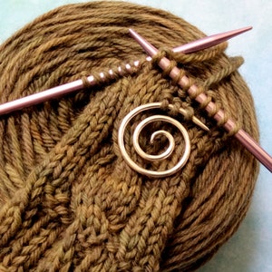 May include: A close-up of a brown yarn ball with two knitting needles and a gold spiral brooch. The yarn is being knit into a brown knitted fabric.