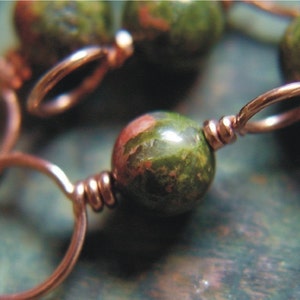 May include: A close-up of a copper wire wrapped around a green and brown speckled stone bead. The bead is part of a necklace or bracelet.
