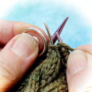 May include: Close-up of a person's hands knitting with a silver knitting ring and purple knitting needles. The yarn is a dark green color.