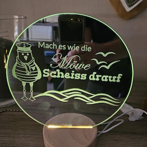 Könnte beinhalten: Ein rundes, klares Acrylschild mit grünem Leuchten. Das Schild zeigt eine Karikatur einer Möwe mit Matrosenhut und dem Text "Mach es wie die Möwe Scheiss drauf". Das Schild steht auf einem Holzsockel.
