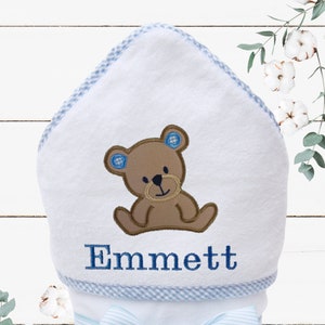 May include: White hooded towel with blue and white checkered trim, a brown bear applique, and the name "Emmett" embroidered in blue. The towel is tied with a blue and white striped ribbon.