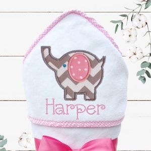 May include: White hooded baby towel with a pink and gray elephant applique. The towel is wrapped and tied with a bright pink satin ribbon. The name "Harper" is embroidered in pink.