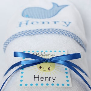 May include: A white towel with a blue whale and the name "Henry" embroidered on it. The towel is rolled up and tied with a blue ribbon. A white tag with a yellow sun and the text "Welcome Henry" is attached to the ribbon.