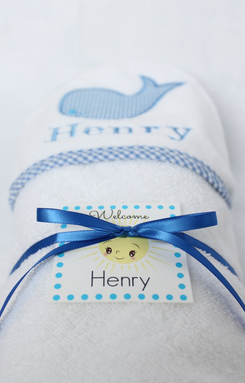 May include: A white towel with a blue whale and the name "Henry" embroidered on it. The towel is rolled up and tied with a blue ribbon. A white tag with a yellow sun and the text "Welcome Henry" is attached to the ribbon.