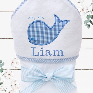 May include: White hooded towel with a blue and white gingham whale design and the name "Liam" embroidered on the bottom. The towel is tied with a blue and white striped ribbon.