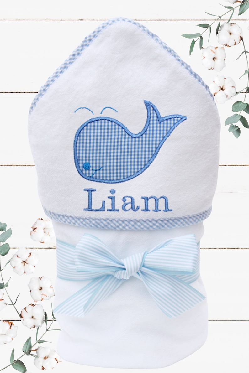 May include: White hooded towel with a blue and white gingham whale design and the name "Liam" embroidered on the bottom. The towel is tied with a blue and white striped ribbon.
