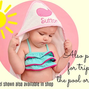 May include: A white hooded baby towel with a pink whale design and the name "Sutton" embroidered on it. The towel has a pink trim. The baby is wearing a turquoise swimsuit with pink trim. A yellow sun graphic is in the background.