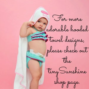 May include: A baby wearing a teal and pink ruffled swimsuit and a white hooded towel with pink trim. The towel is personalized with the name "Sutton". The background is a solid pink.