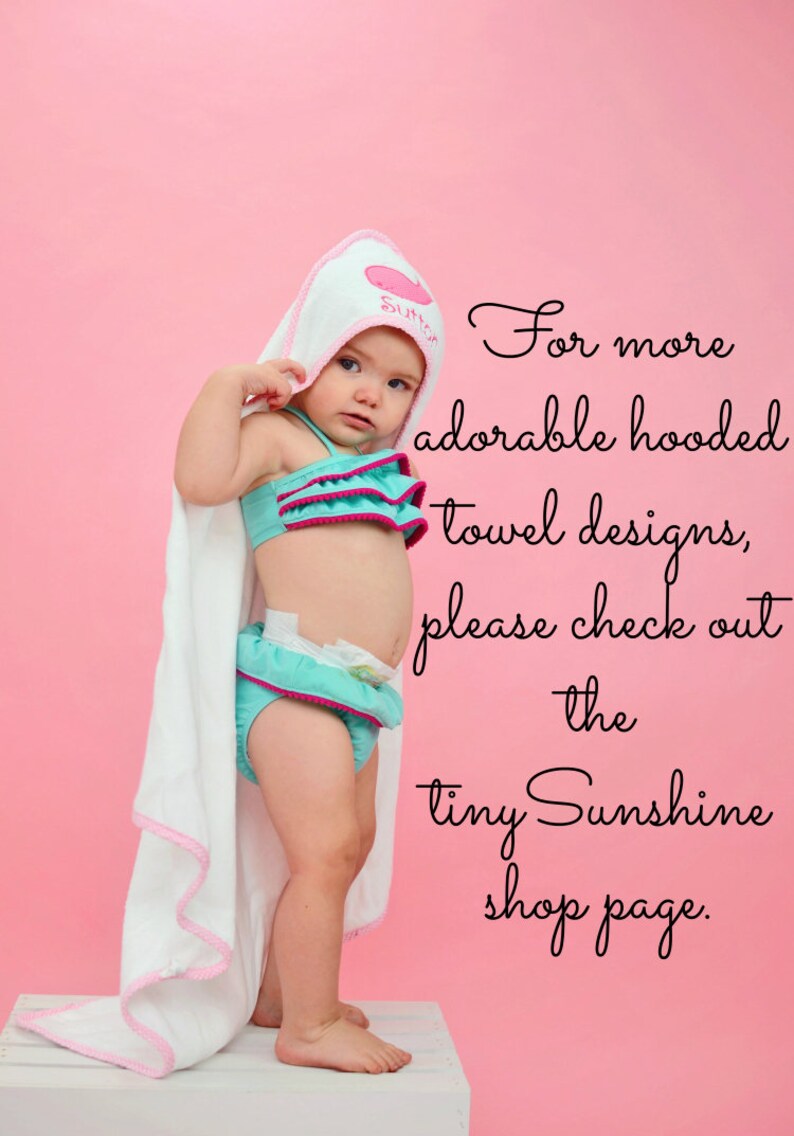 May include: A baby wearing a teal and pink ruffled swimsuit and a white hooded towel with pink trim. The towel is personalized with the name "Sutton". The background is a solid pink.